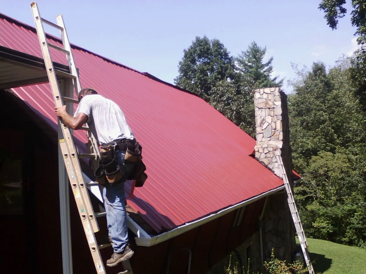 Roofer on a ladder beside a red metal roof and stone chimney — Soffit & Fascia Repair by Roofing Brothers in Ordway